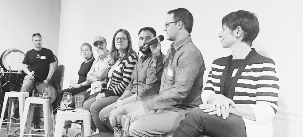 Andrew speaking on a microphone while sitting on a stool with a host and five other panelist.
