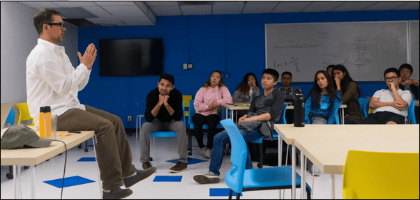 Andrew sitting on a desk speaking to a classroom of students at UCSD